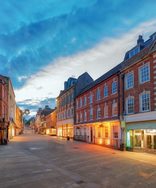 An image of a high street in an unknown town, it's twilight and the shop entrances are lit