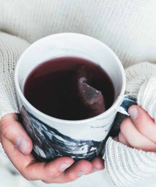 Hands in a white oversized sweater hold a mug of herbal tea that looks like Roobush. The teabag is still in the tea