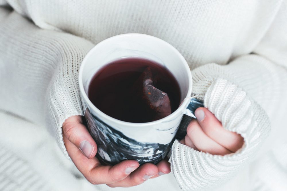 Hands in a white oversized sweater hold a mug of herbal tea that looks like Roobush. The teabag is still in the tea