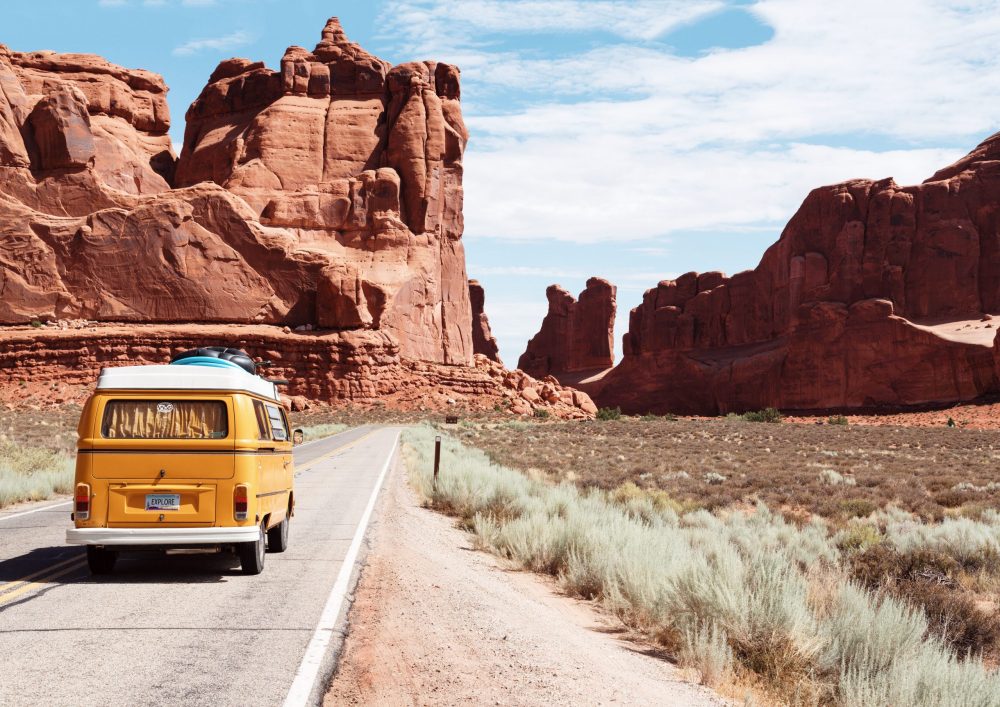 A yellow VW camper van drives along a deserted road towards red coloured mountains