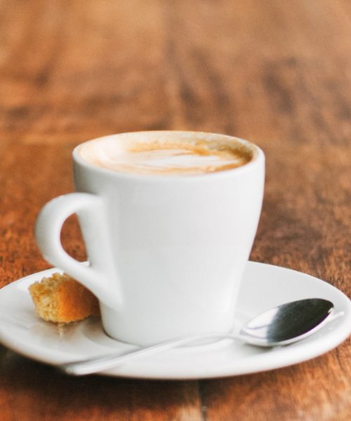 A cup of cappuccino with a white saucer on a wooden counter top